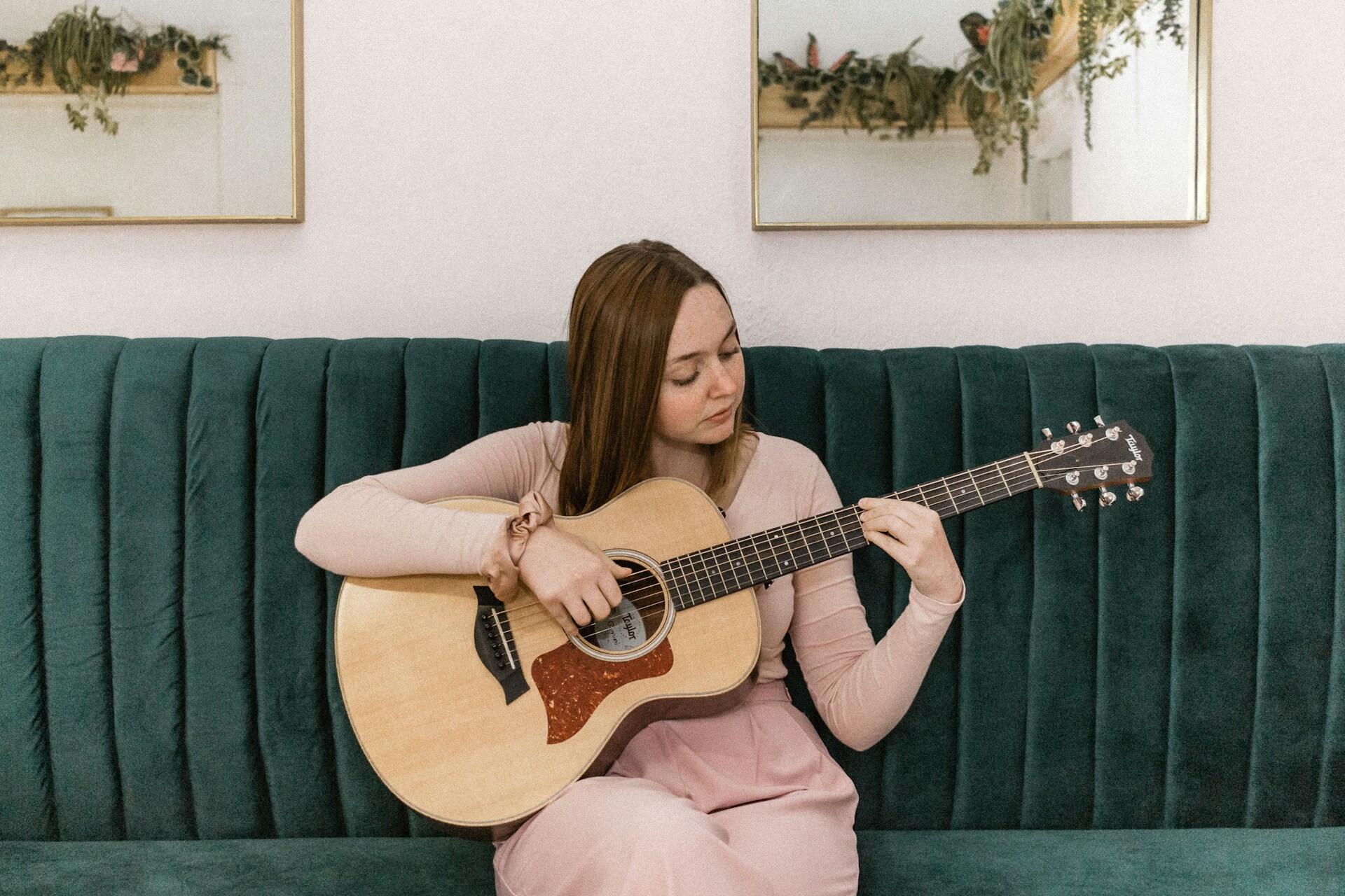 Tenåringsjente sitter på en grønn sofa og øver på akustisk gitar i et rolig hjemmemiljø.