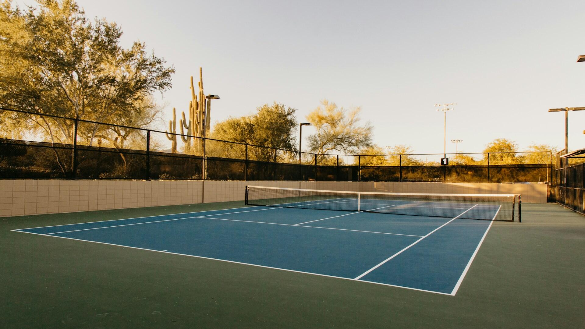 Blå tennisbane i solnedgang med trær og gjerde i bakgrunnen.