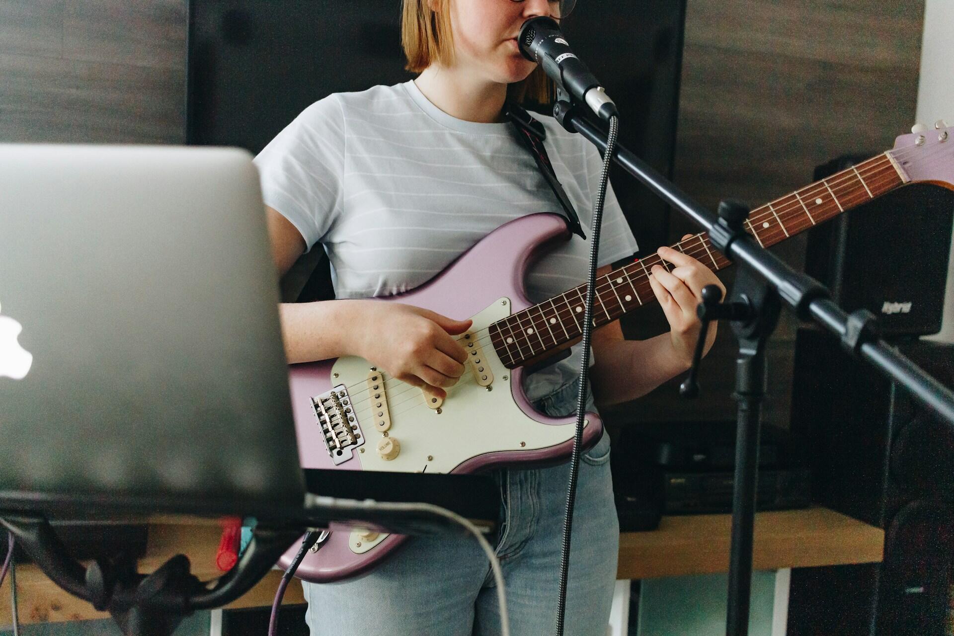 Kvinne med lilla elgitar synger i mikrofon mens hun spiller foran en laptop hjemme.