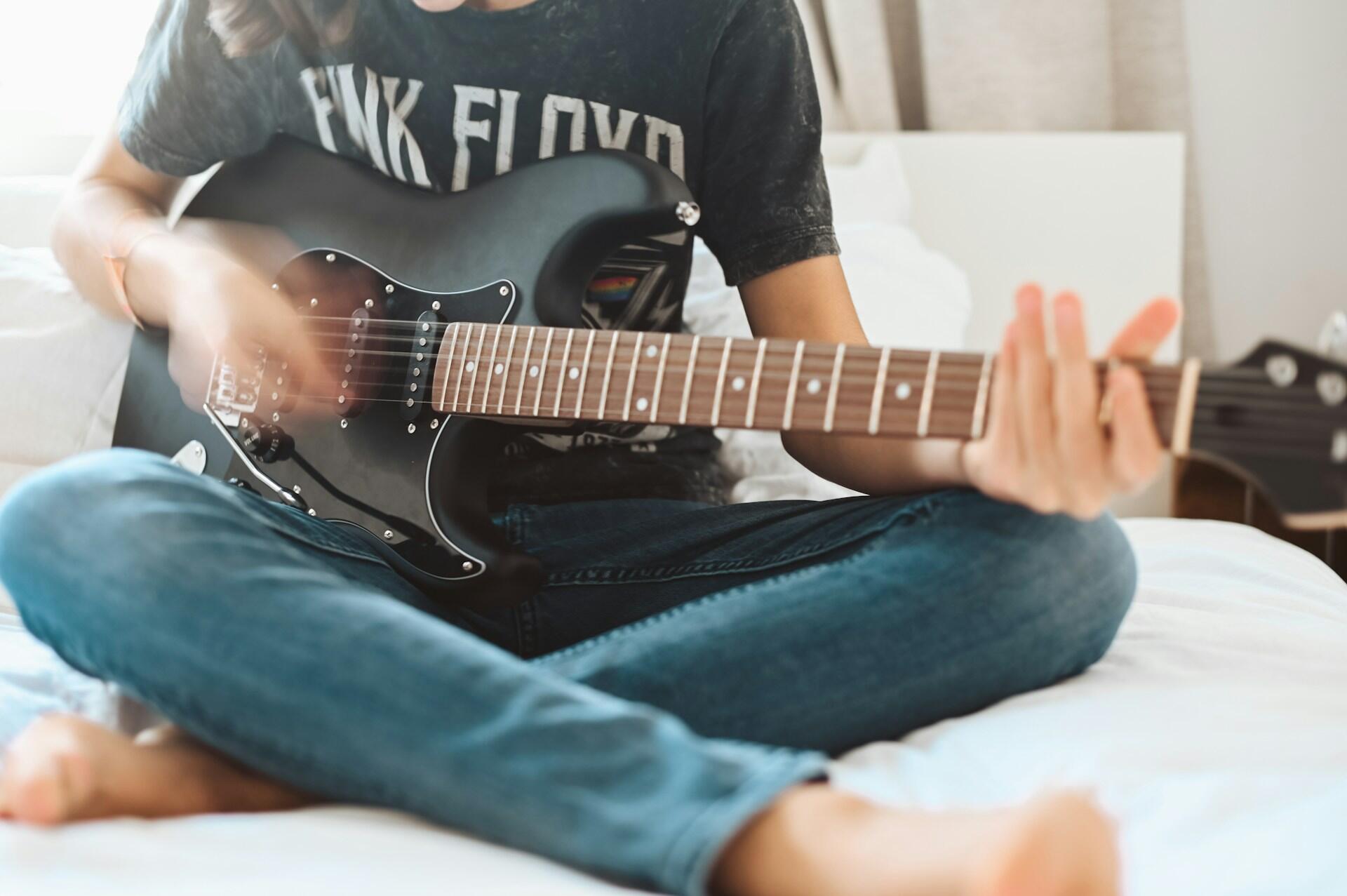Ung person med Pink Floyd-skjorte spiller elgitar mens han sitter i sengen.