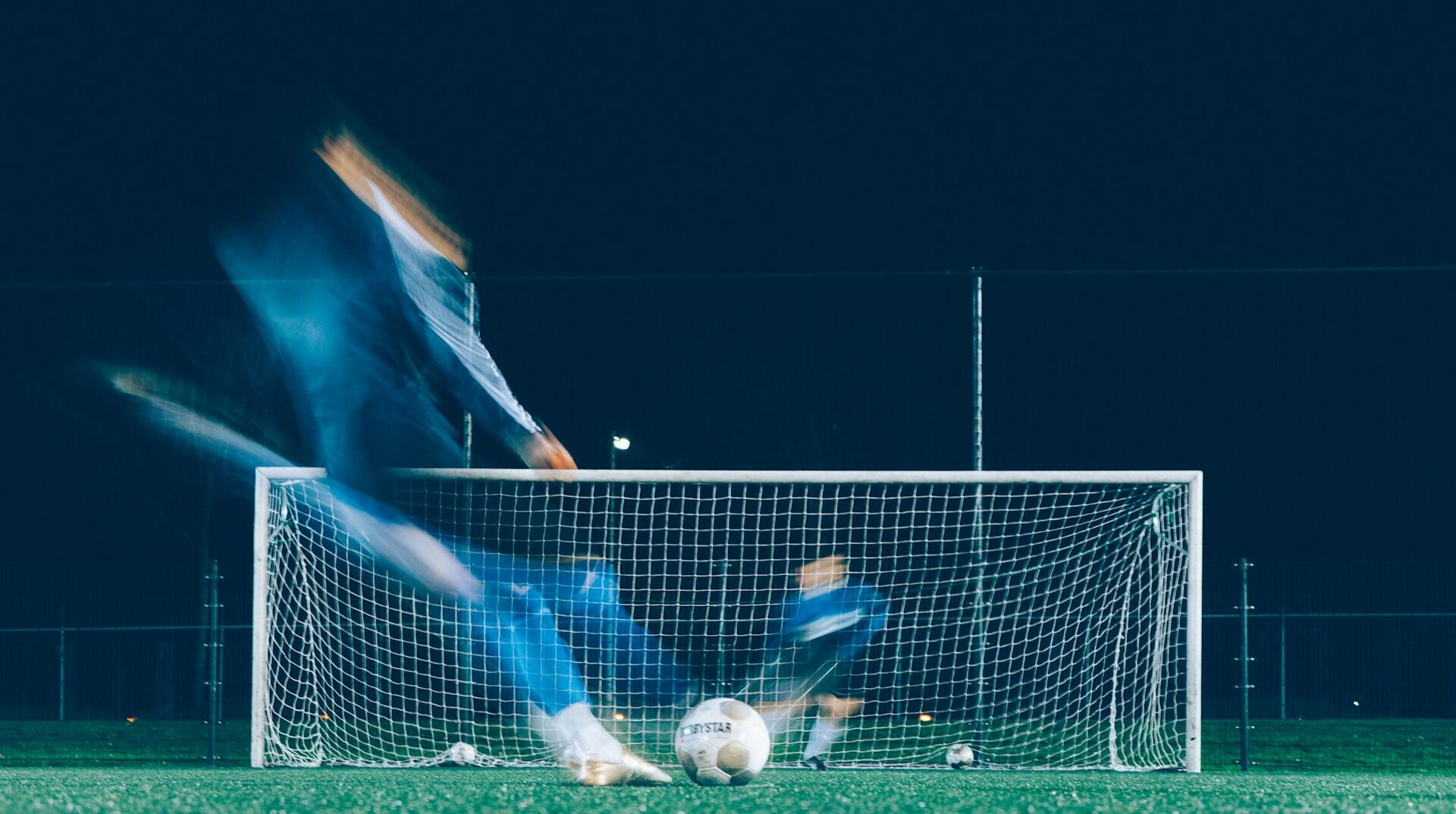 To fotballspillere i blått spiller mot hverandre, en i mål og den andre skal til å sparke.