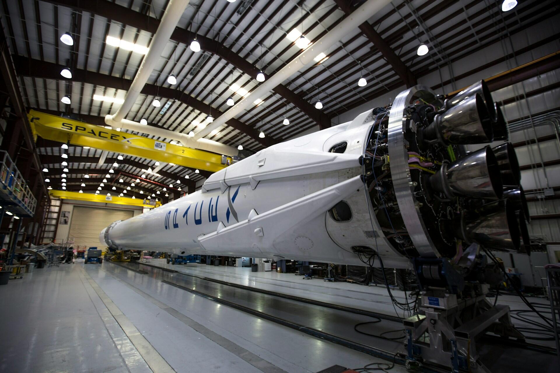 SpaceX sin hvite Falcon-rakett i produksjonshall med gul kran og industrielt miljø