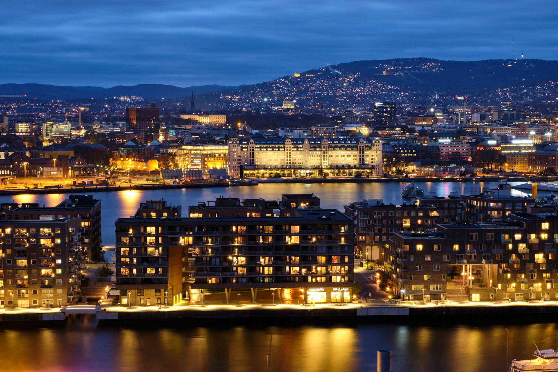 Kveldsbilde av Oslo sentrum med opplyste bygg og fjell i bakgrunnen – symbol på velstand og velferd.
