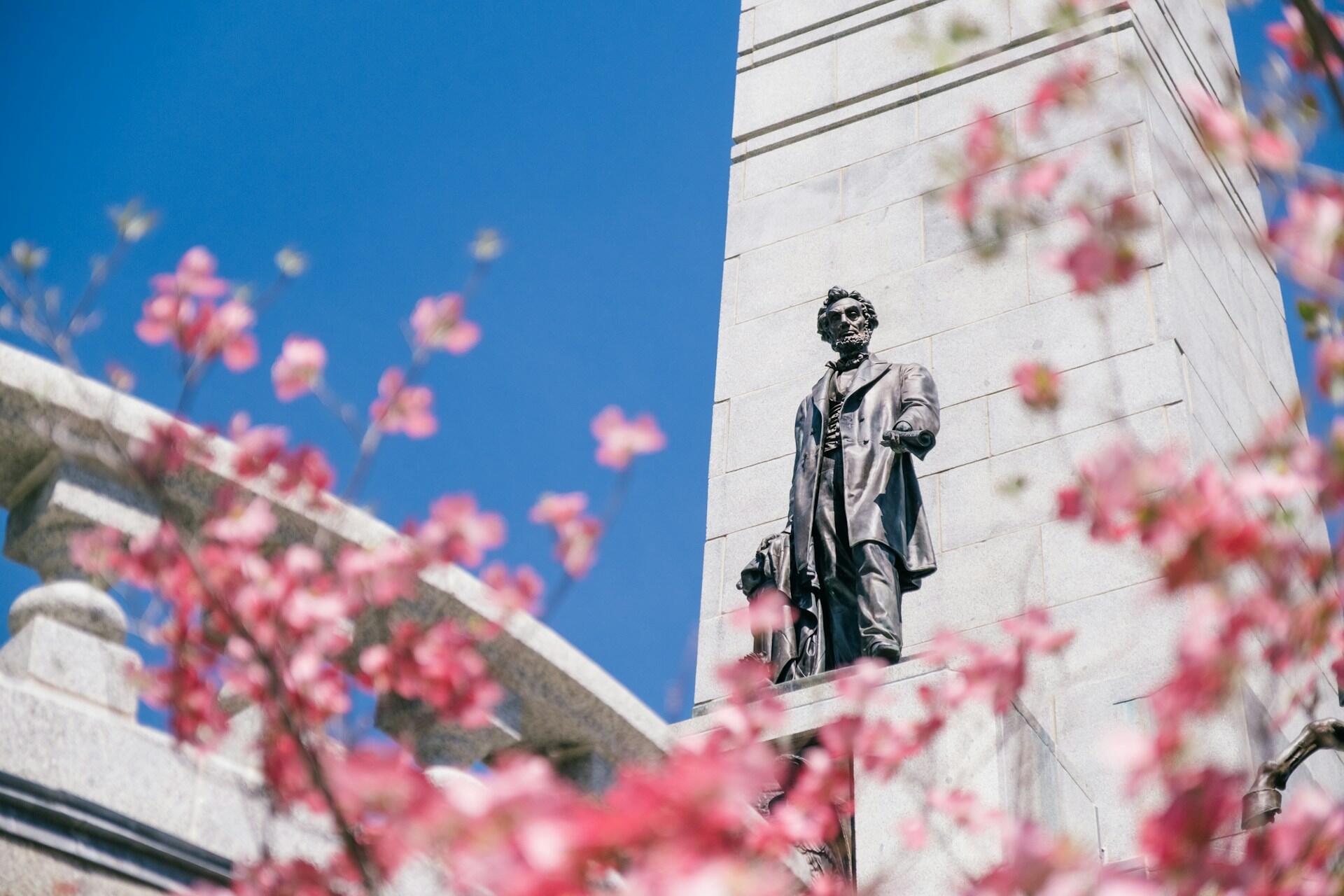 Bronsestatue av Abraham Lincoln foran en granittsøyle, med rosa blomster i forgrunnen og blå himmel.