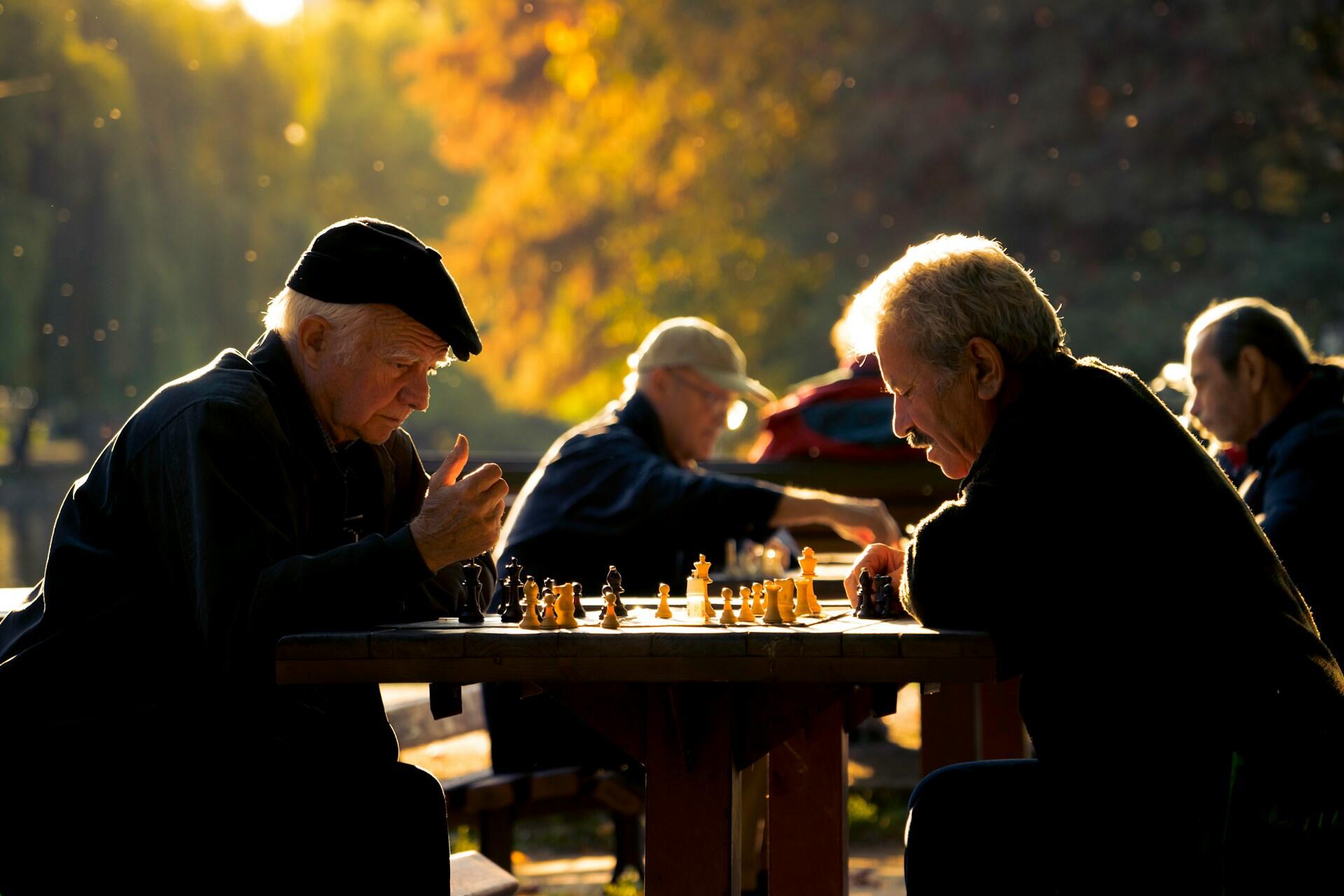 To eldre menn sitter sammen med andre på piknikbord i parken og spiller sjakk