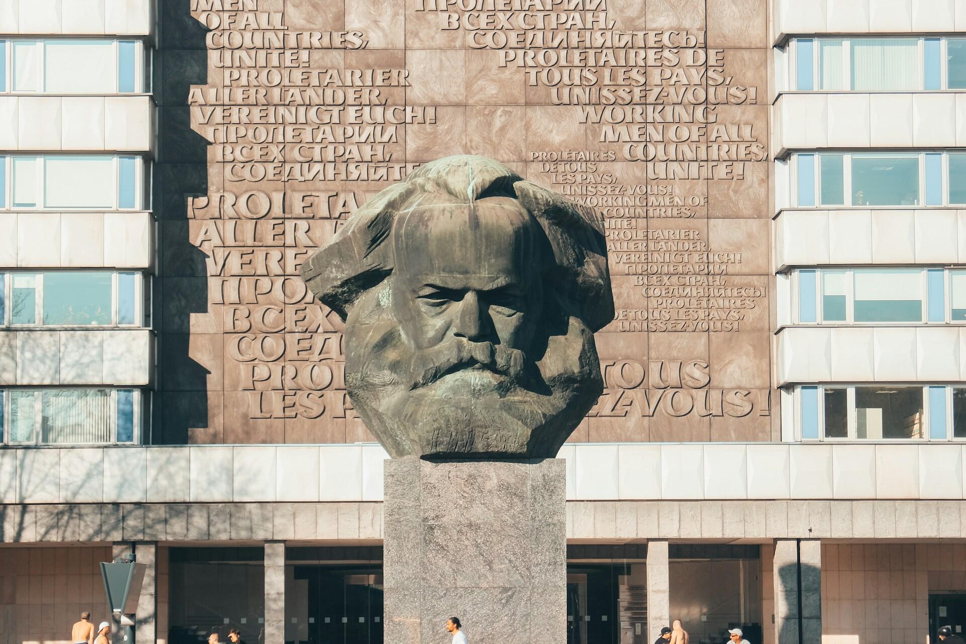 Stor bronsebyste av Karl Marx på sokkel foran en vegg med sitater på flere språk, inkludert «Workers of all countries, unite!»