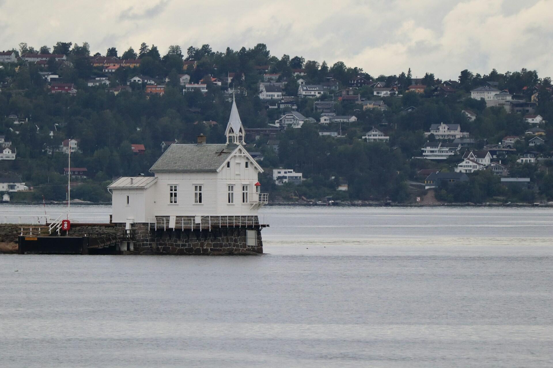 Hvit norsk kirke med spir bygd ut på en kai over fjorden, med bebyggelse i åsene i bakgrunnen