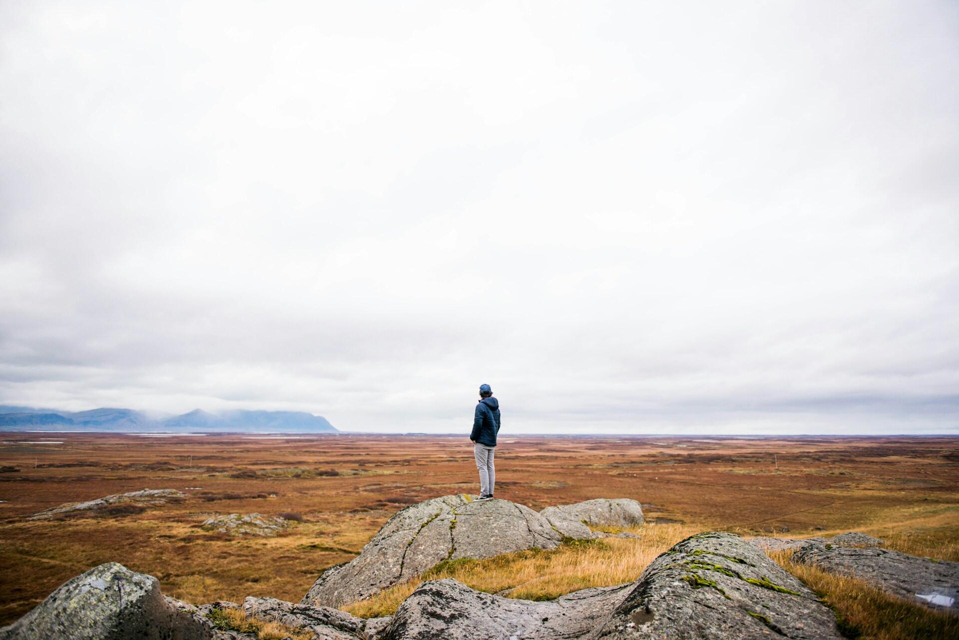 Person står alene på en stor stein og ser utover et åpent, øde landskap med fjell i horisonten