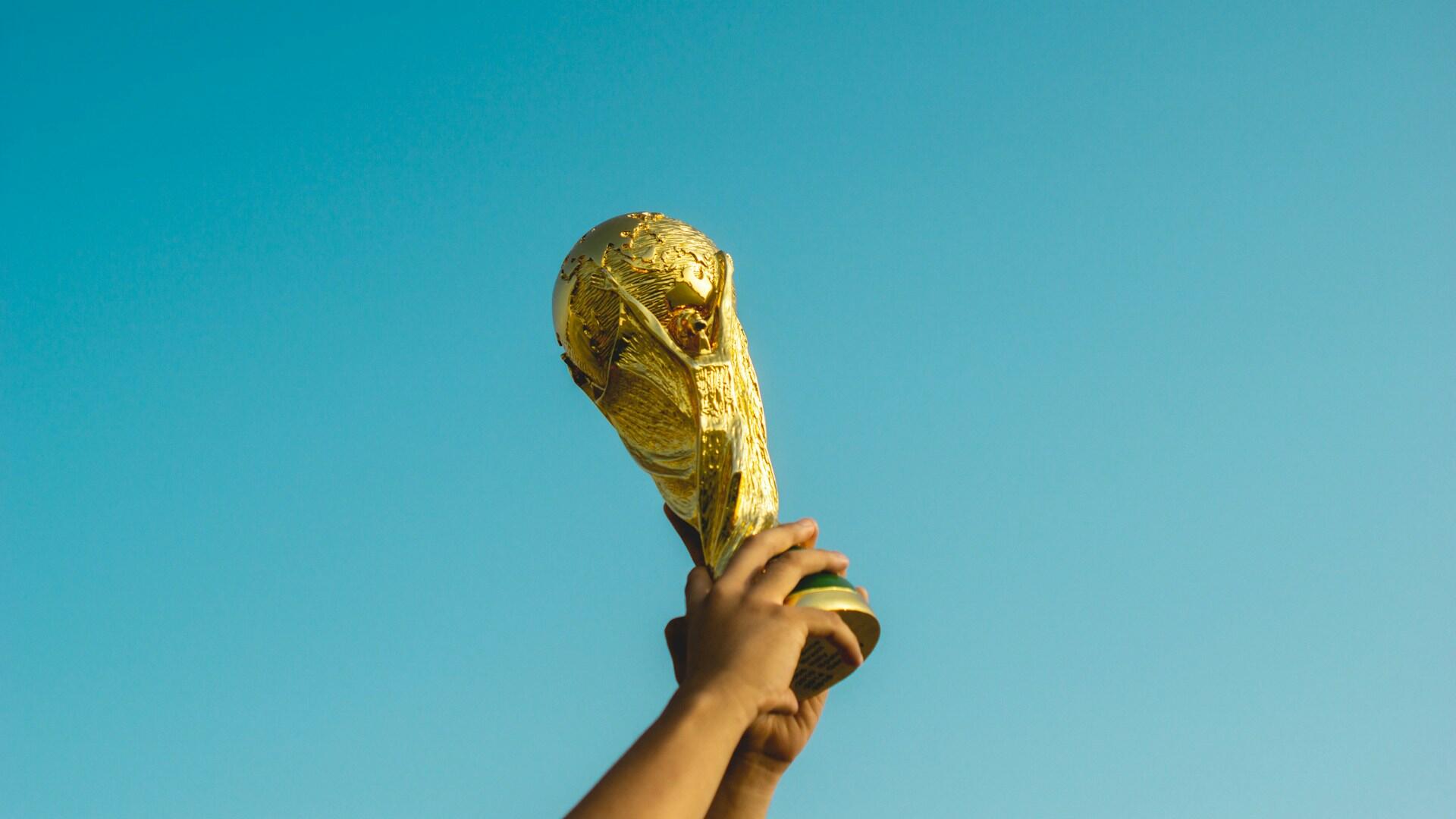 Hender som holder fifa trofeet i lufta med blå himmel bak