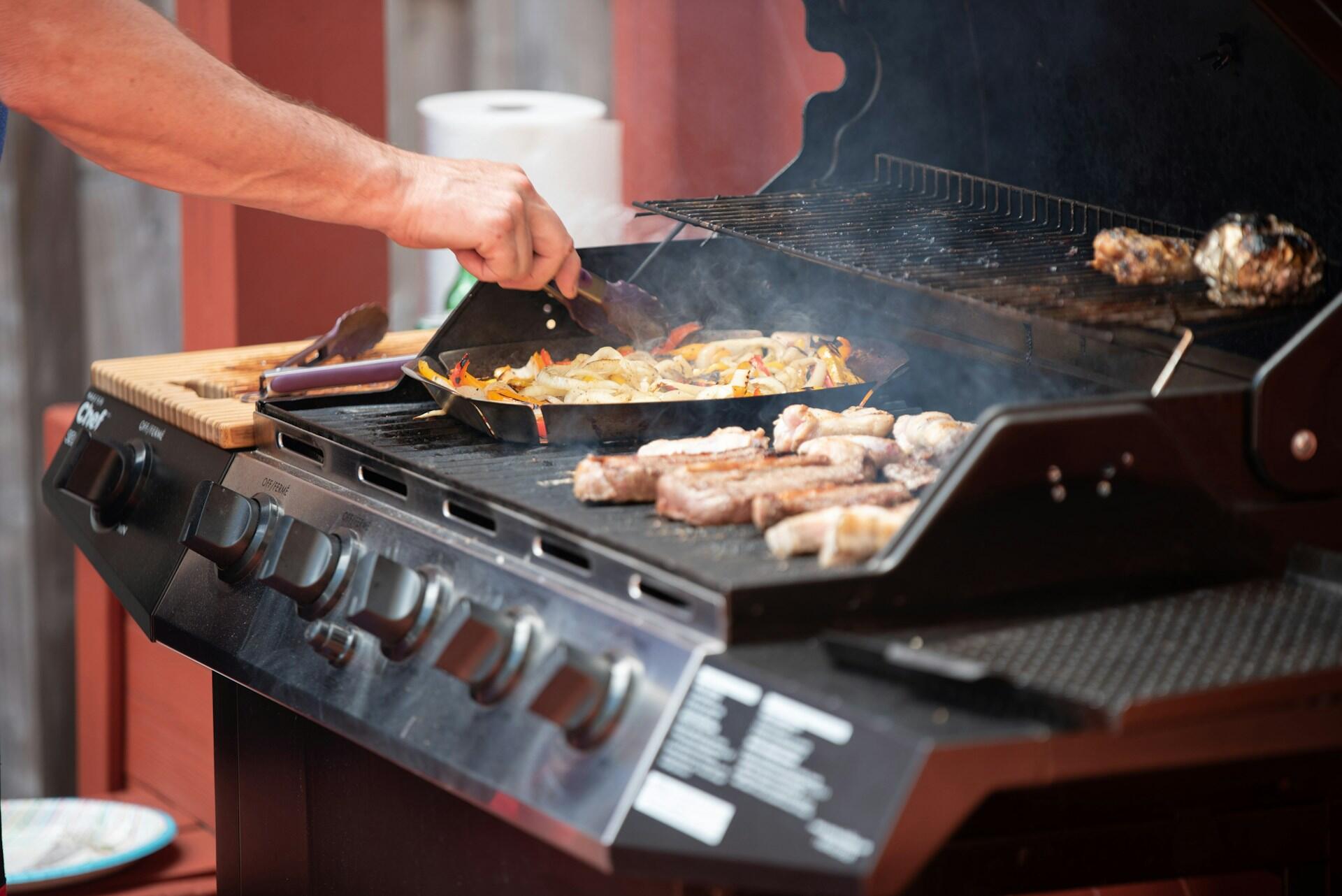 Gassgrill med grønnsaker i grillpanne og kjøtt på risten under matlaging.