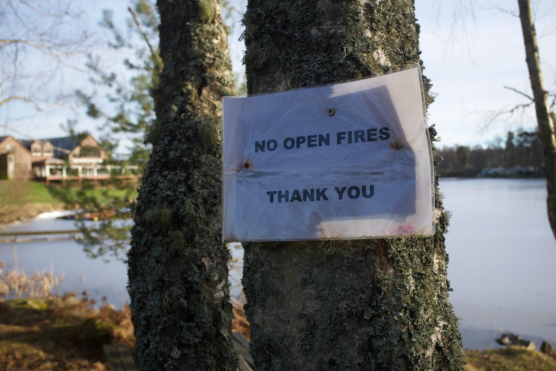 Skilt med teksten «No open fires» festet på et tre ved et vannområde.