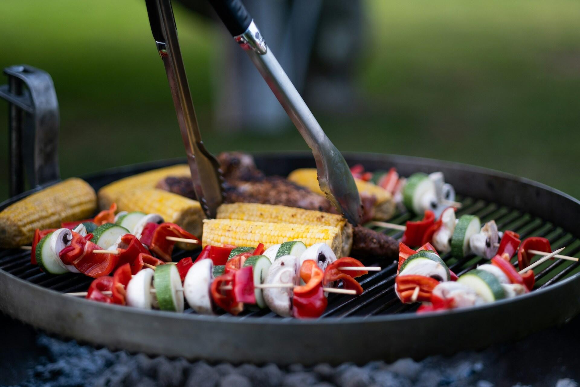 Grønnsaksspyd, maiskolber og kjøtt grilles på kullgrill med grilltang.
