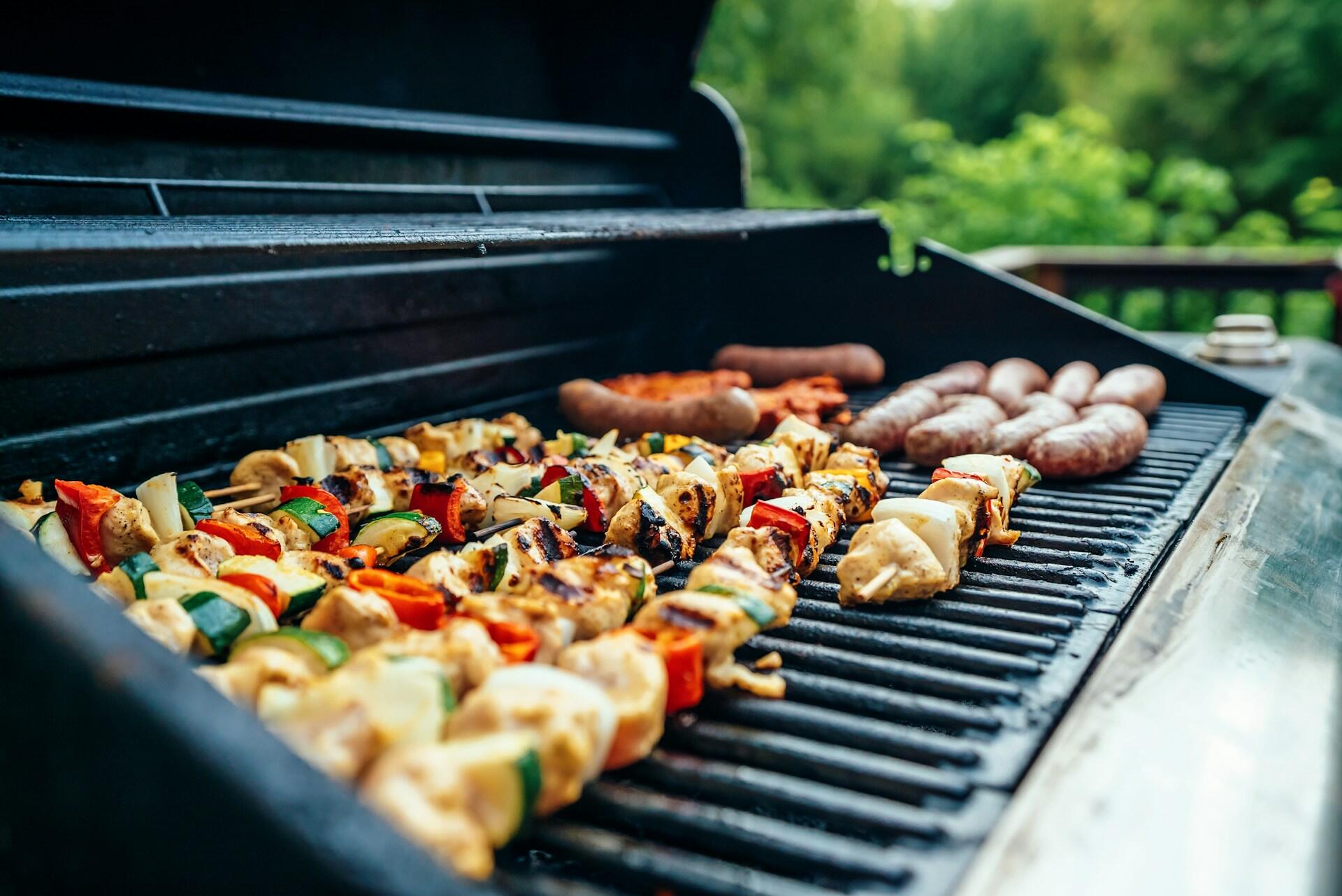 Grillspyd med kylling og grønnsaker grilles sammen med pølser på en stor grill.