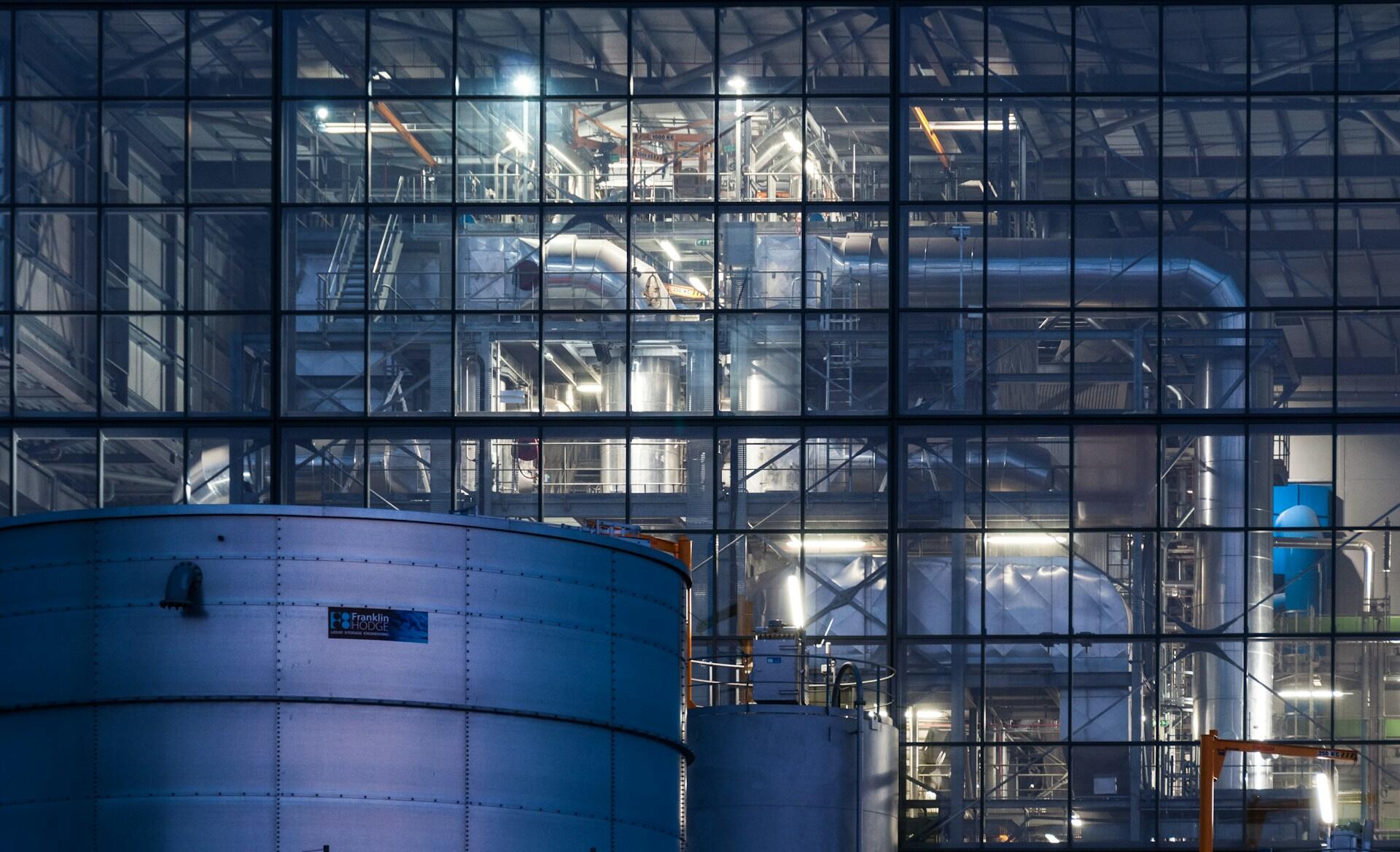 Industrielt prosessanlegg med tanker, rør og produksjonsutstyr bak en glassfasade.