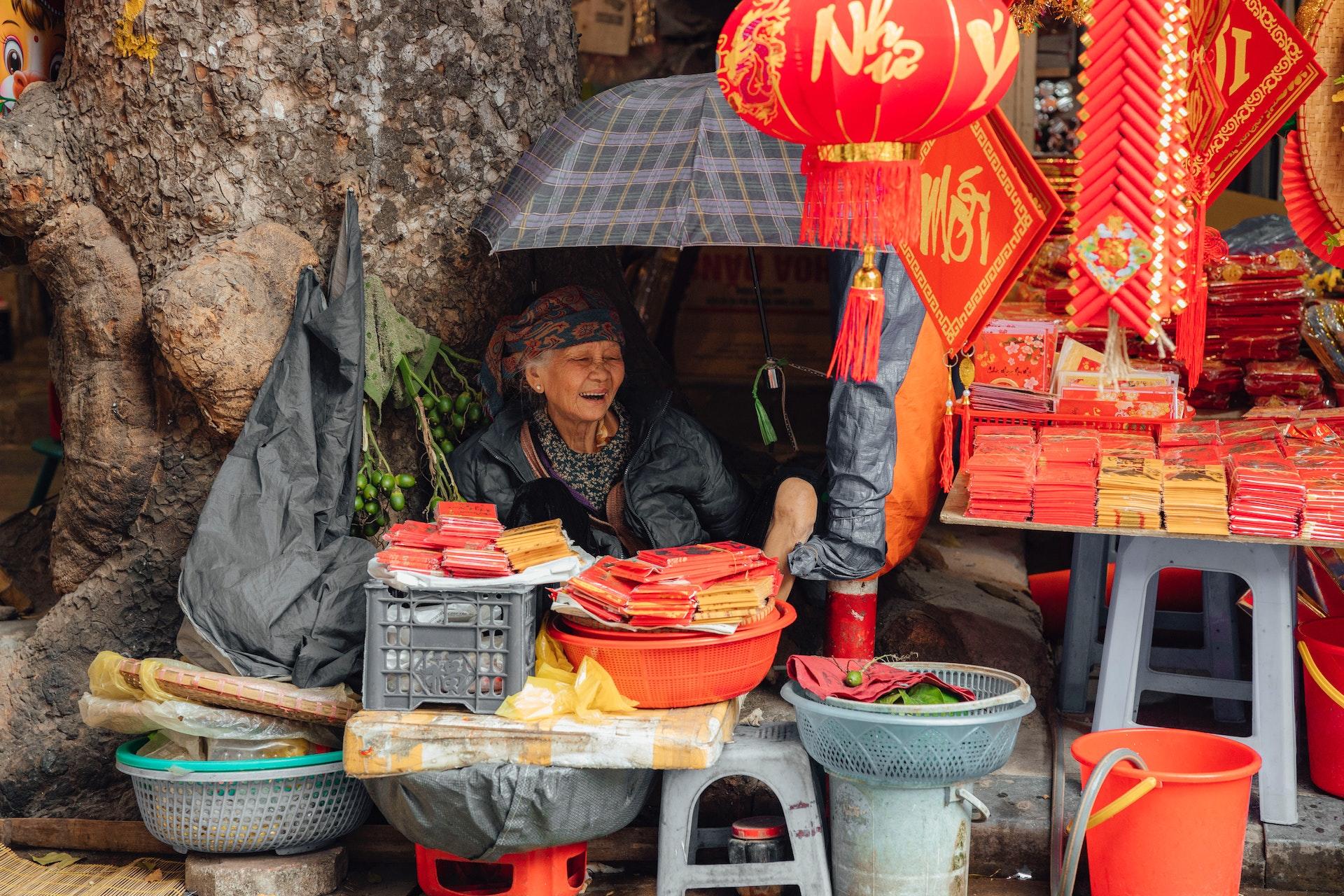 An elderly woman mans a stall selling bright red Chinese New Year decorations.