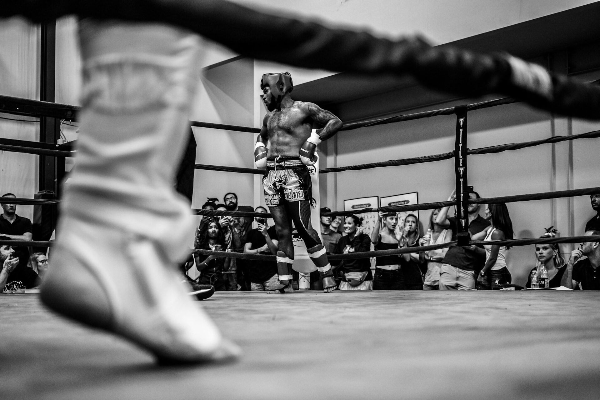 Black-and-white view across a boxing ring with a blurred shoe in the foreground and a fighter near the ropes
