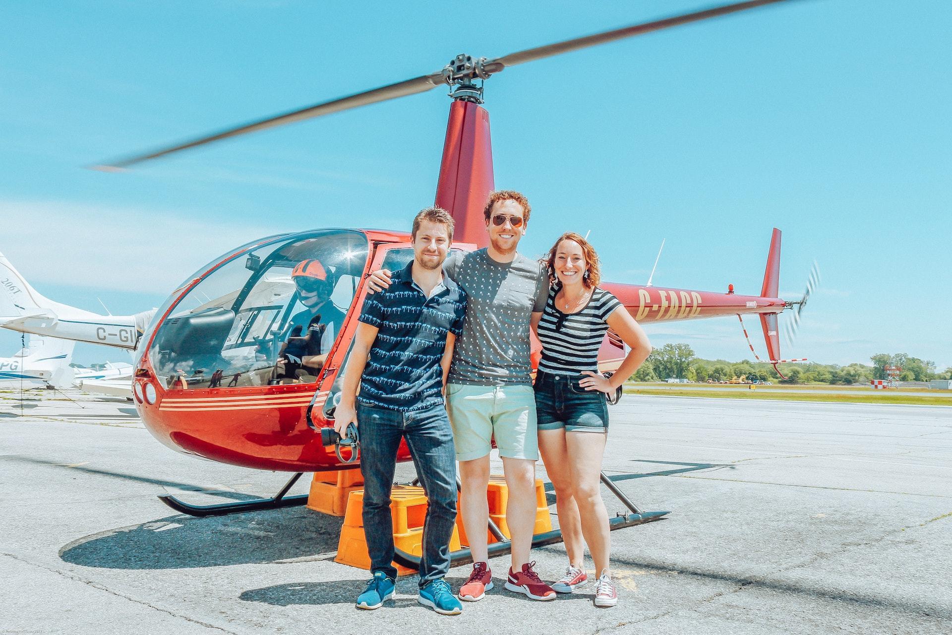 Three people standing in front of a helicopter smiling at the camera on a sunny day.