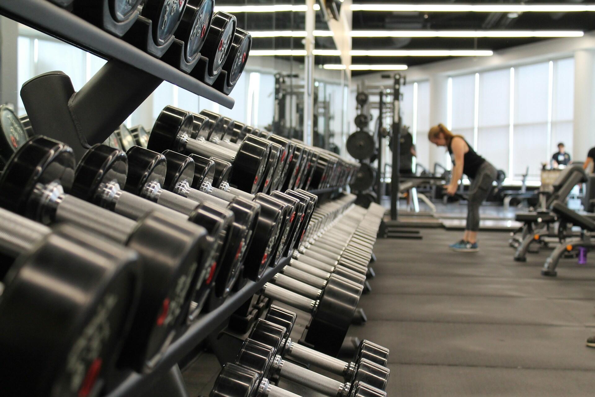 Rows of dumbbells in a bright gym with a woman exercising in the background
