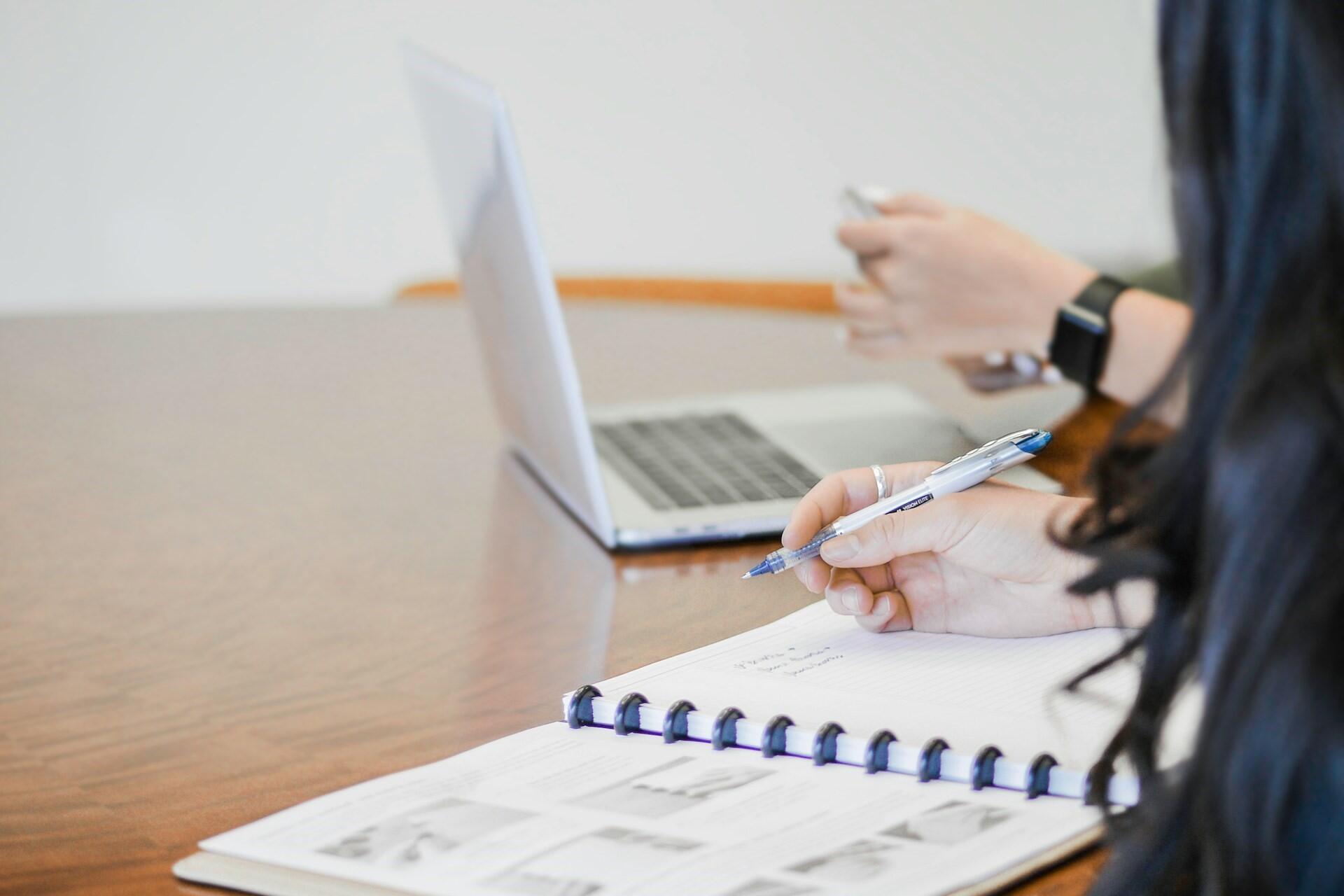 person writing notes in a notebook while working on a laptop