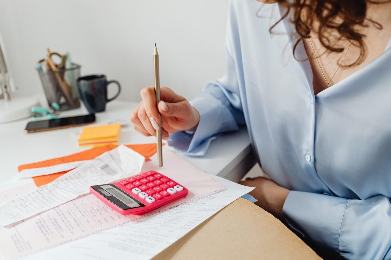 A basic red calculator on a table where a person pokes the number keys with a pencil.