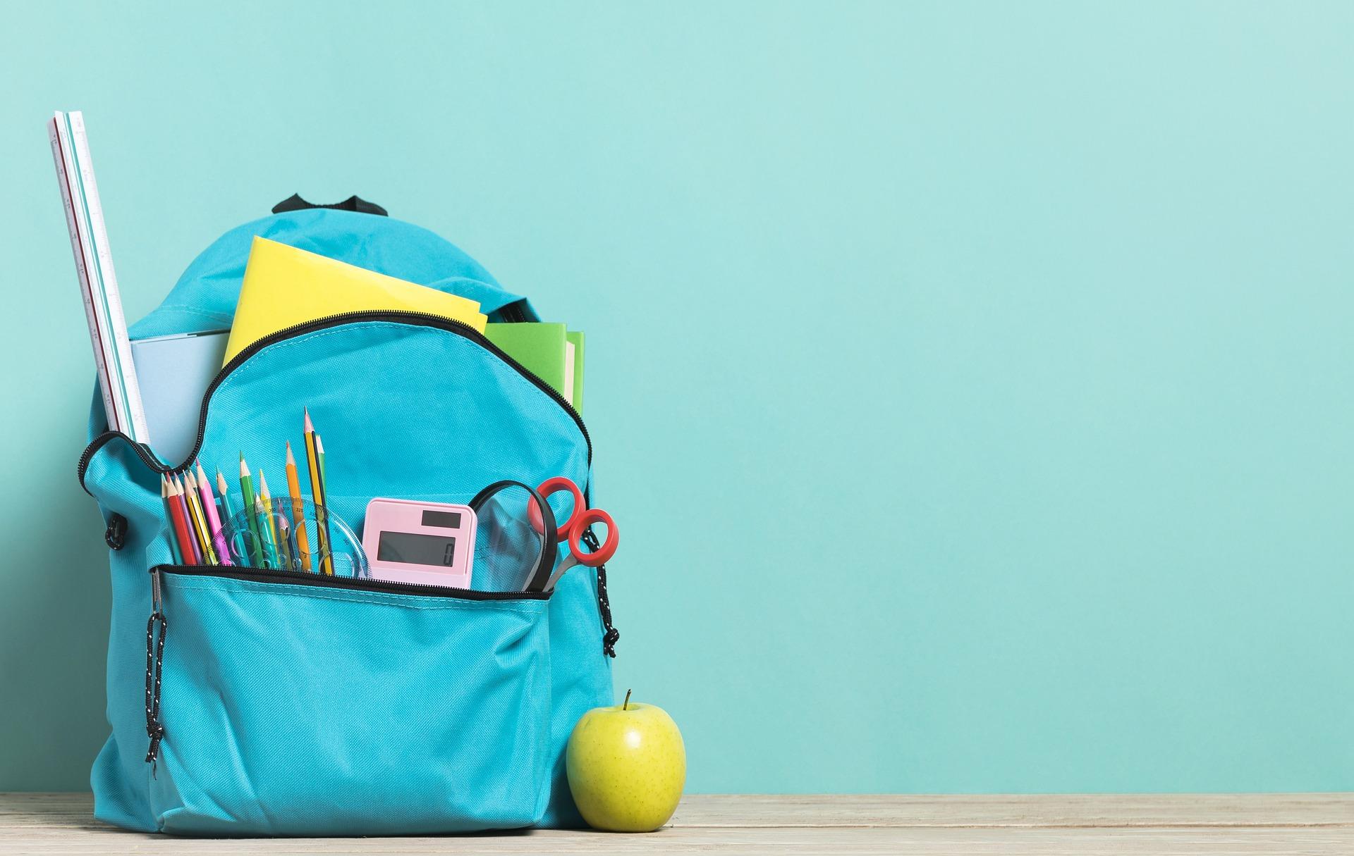 A blue backpack full of school supplies against a lighter blue wall.