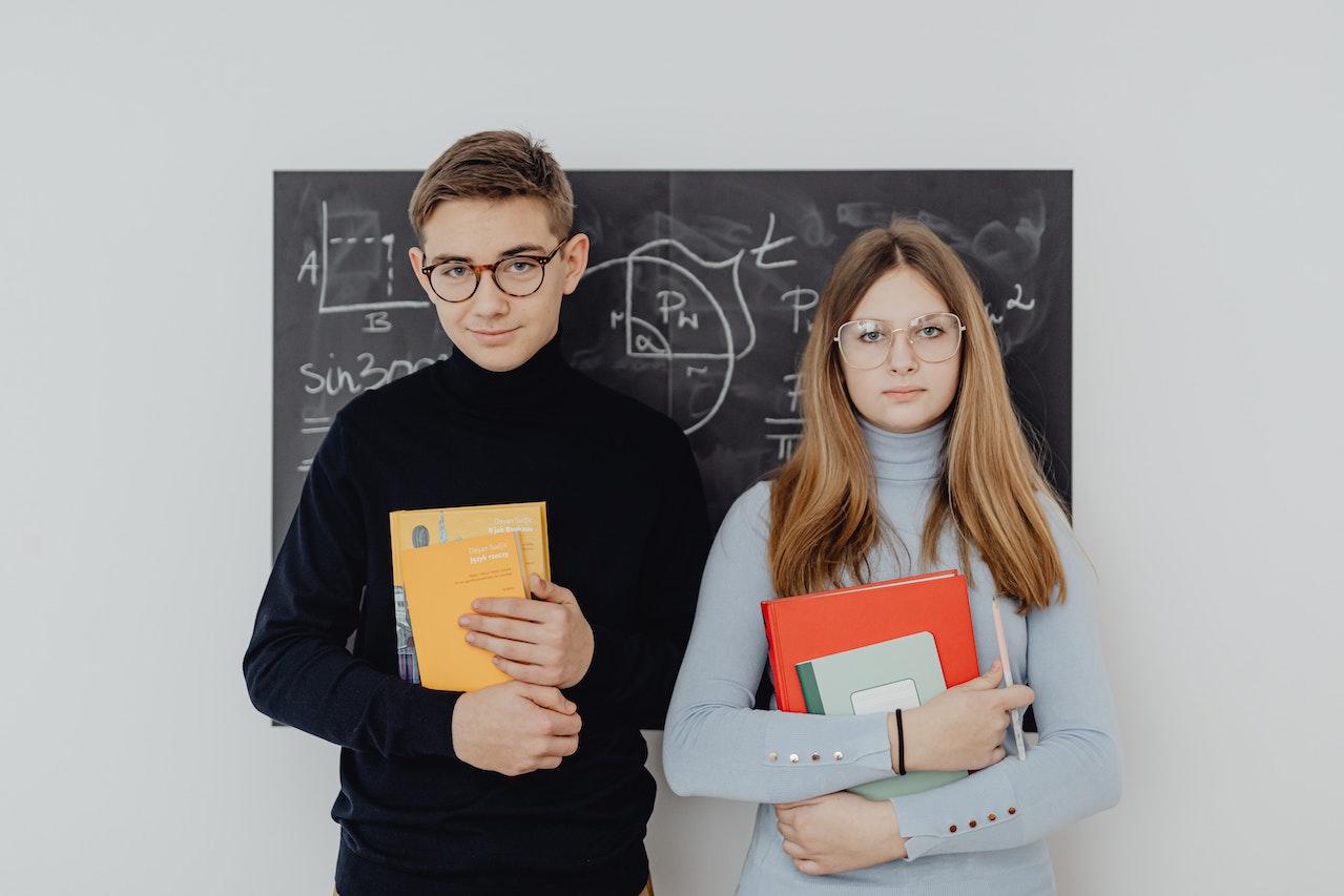 Two students stand in front of a chalkboard holding folders and other school supplies.