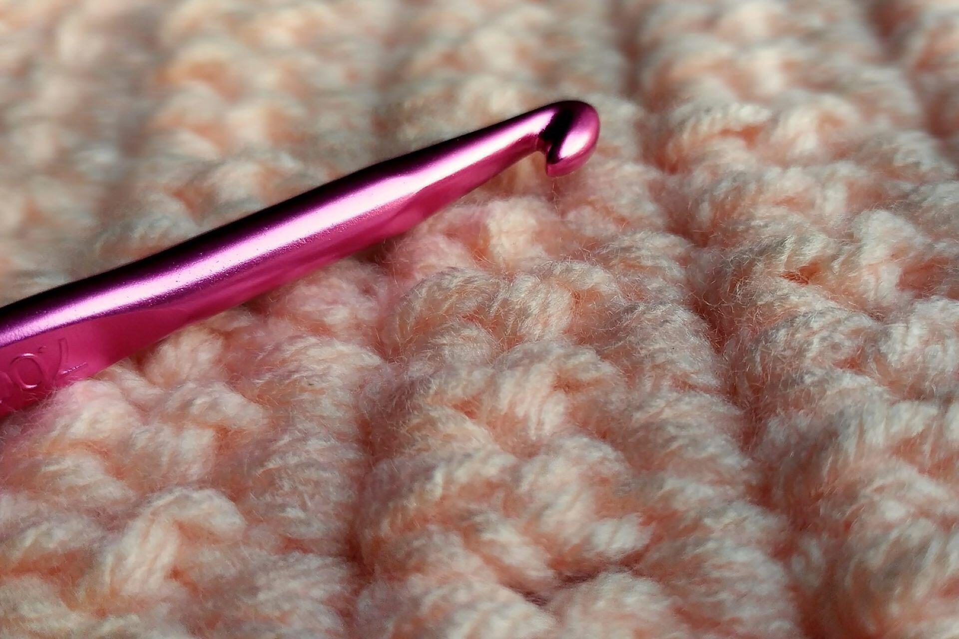 A pink crochet hook resting on pale pink crocheted fabric.