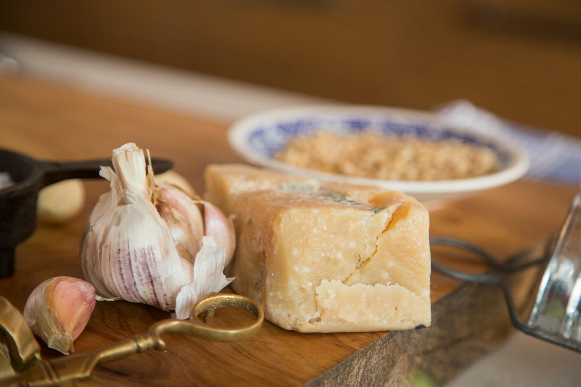 Block of cheese beside garlic cloves on a wooden cutting board, with a small cast-iron pan and a patterned plate in the background.