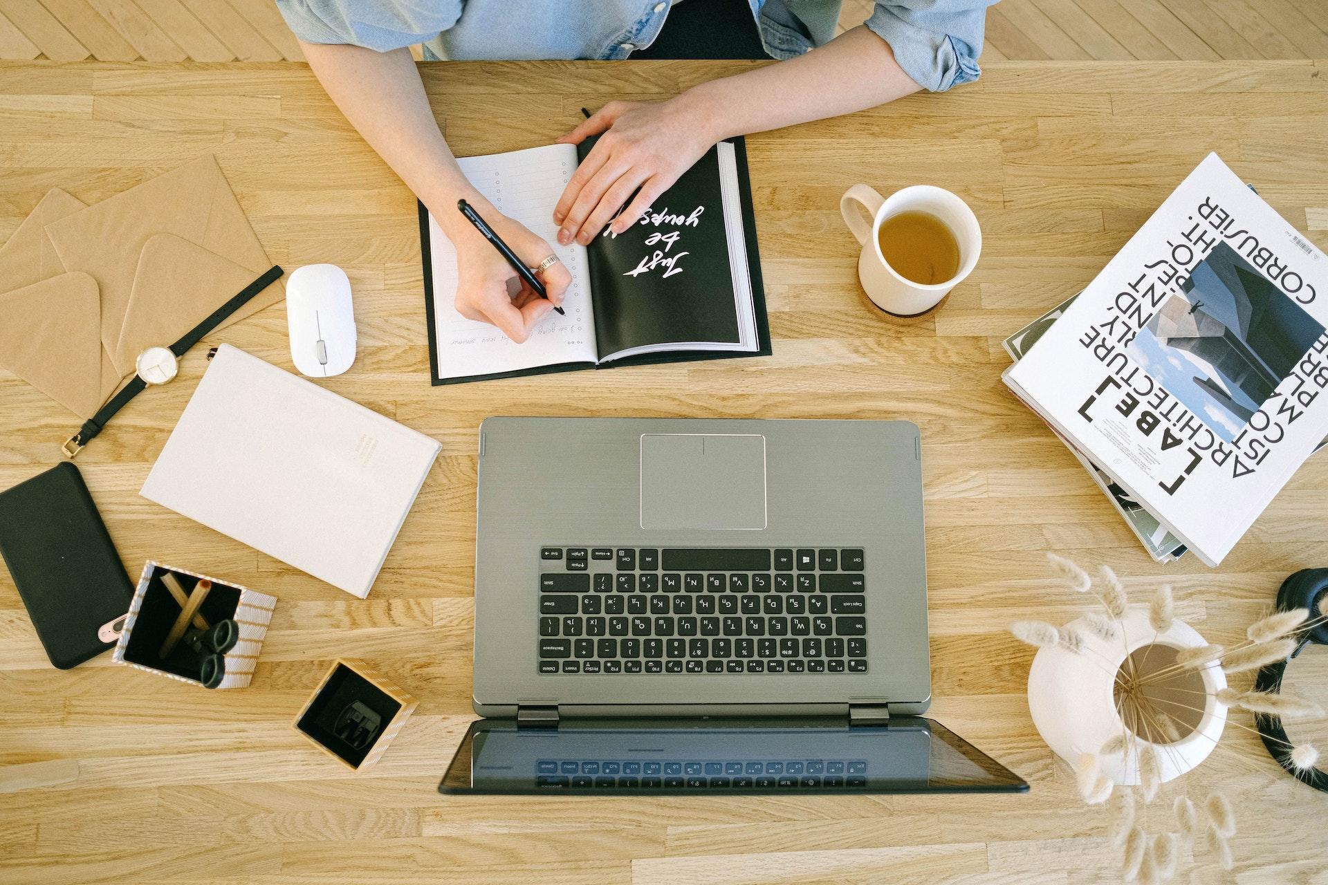 A flat-lay of a person at a desk with a laptop, planner, tea, and other papers nearby.