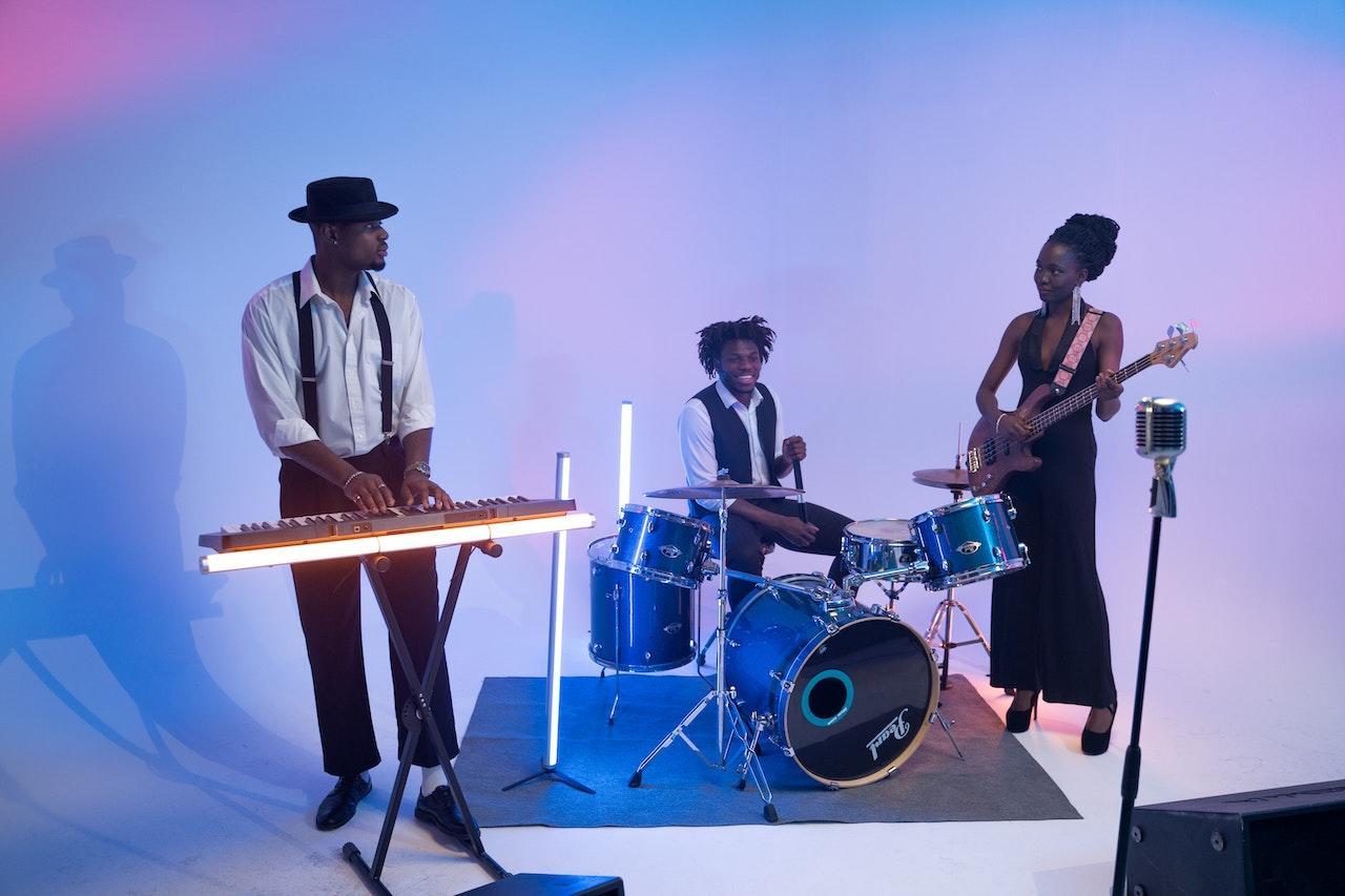 Three band members stand on a white floor with a white background with neon lights for accent colors. There is a keyboardist, a drummer, and a guitarist.