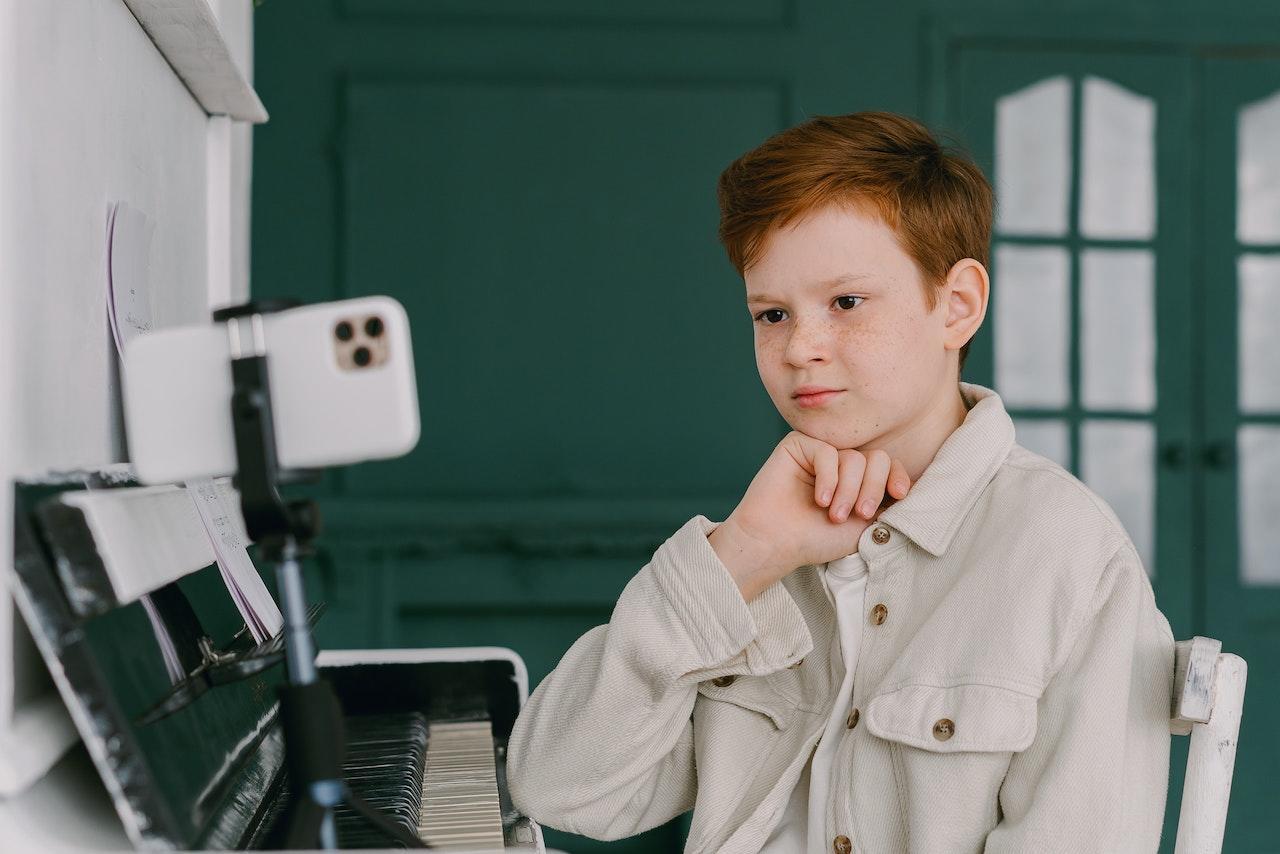 A young piano student sits at his home piano with a smartphone on a stand nearby, seemingly watching instruction on the screen.
