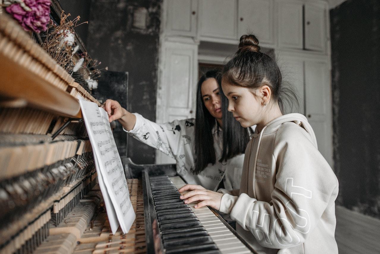 A piano teacher in the background holds open the sheet music book for the young student sitting on the piano bench in the foreground.