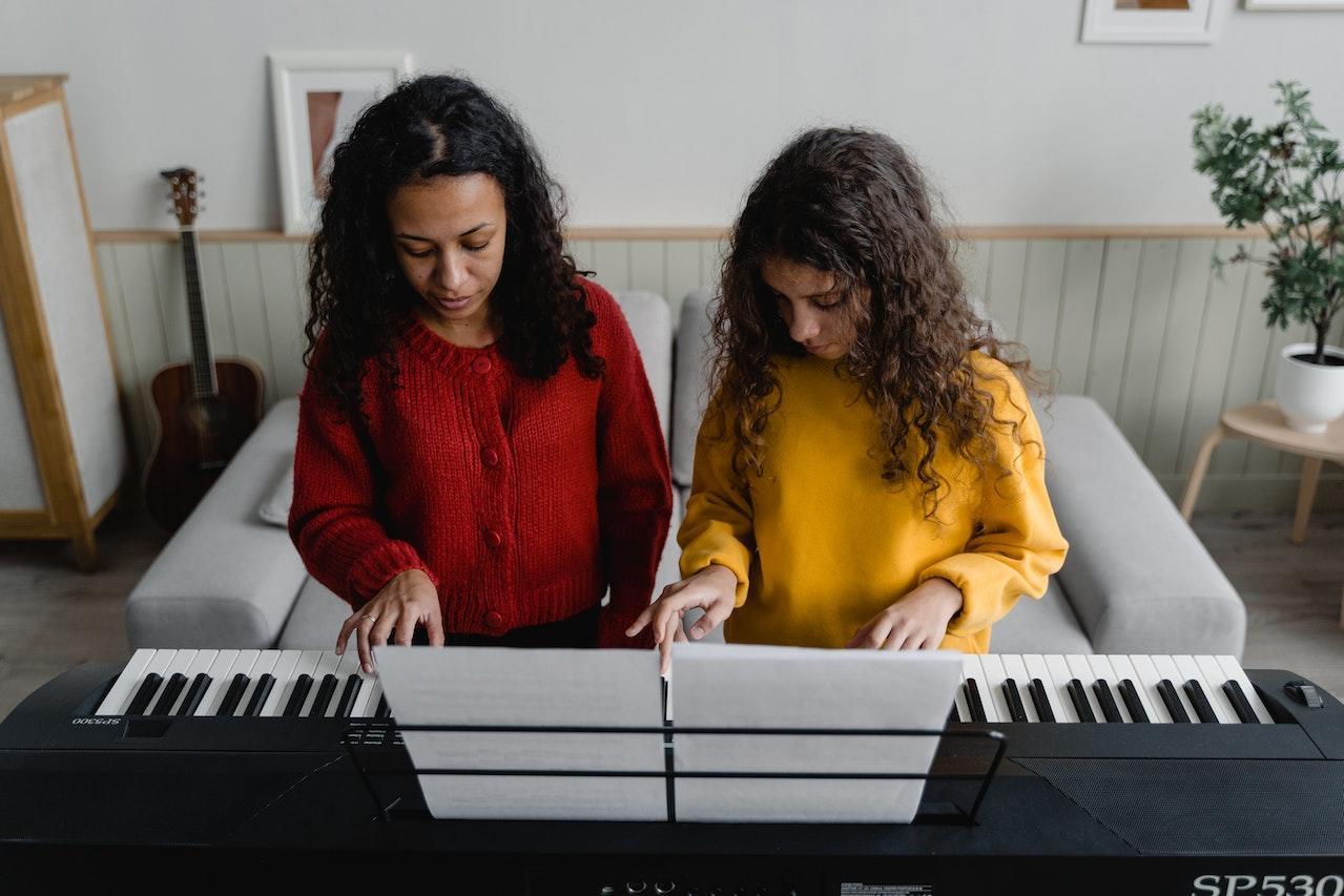 A woman teaches a young girl how to play the piano as they both sit on the piano bench.