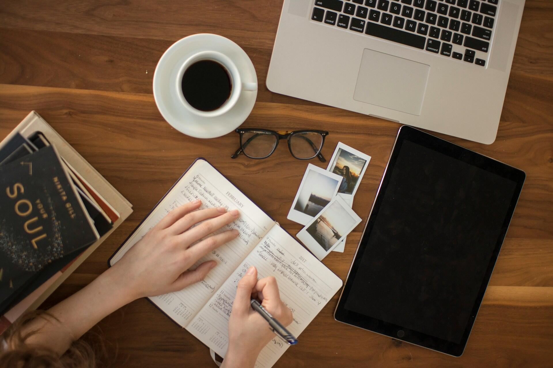 Open planner, laptop, tablet, books, glasses and coffee on a wooden table