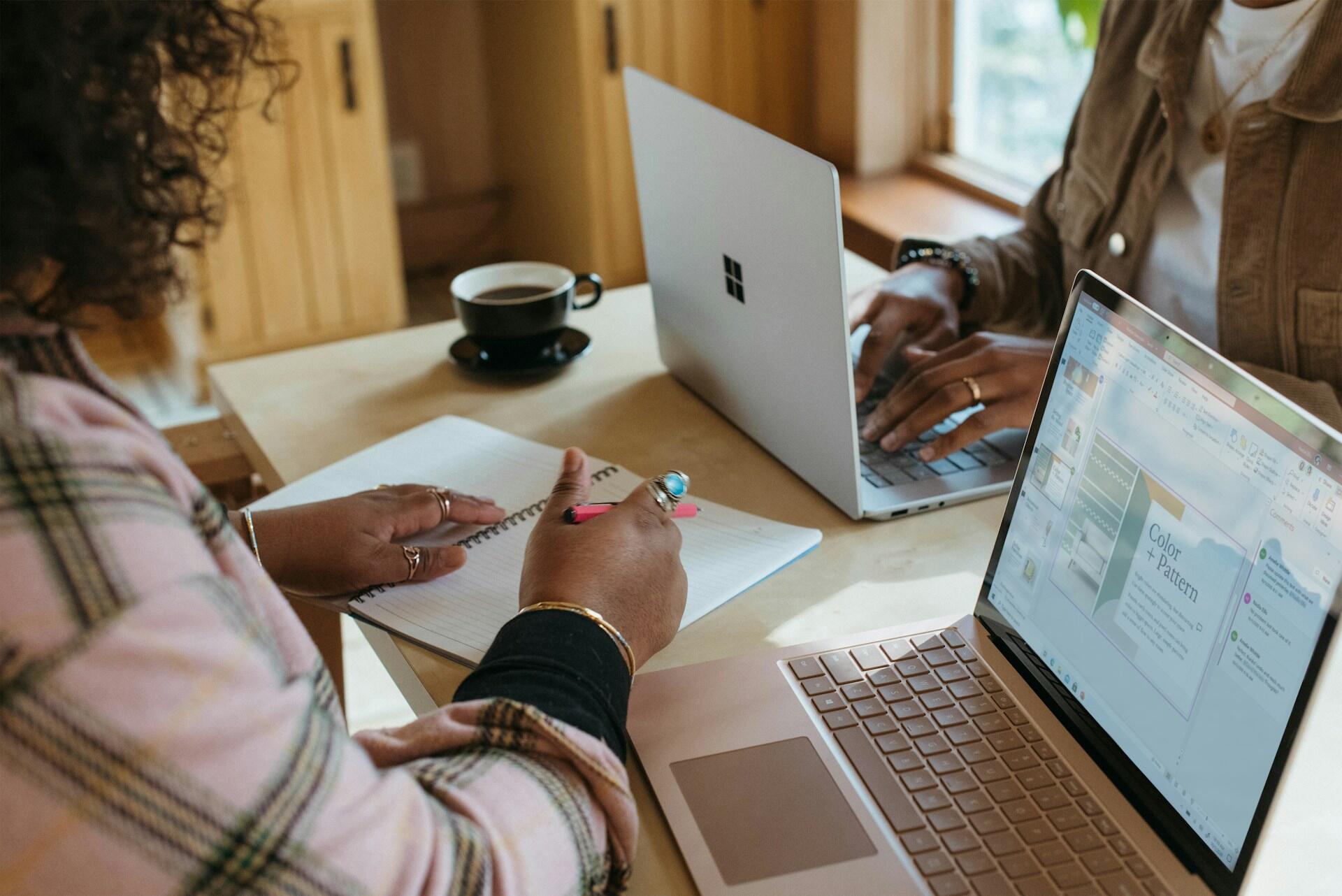 Two people using laptops while one writes in a spiral notebook at a table