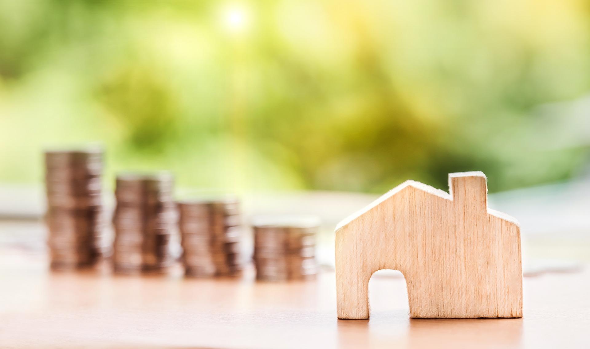 wood cutout of house shape in front of blurred stacks of coins