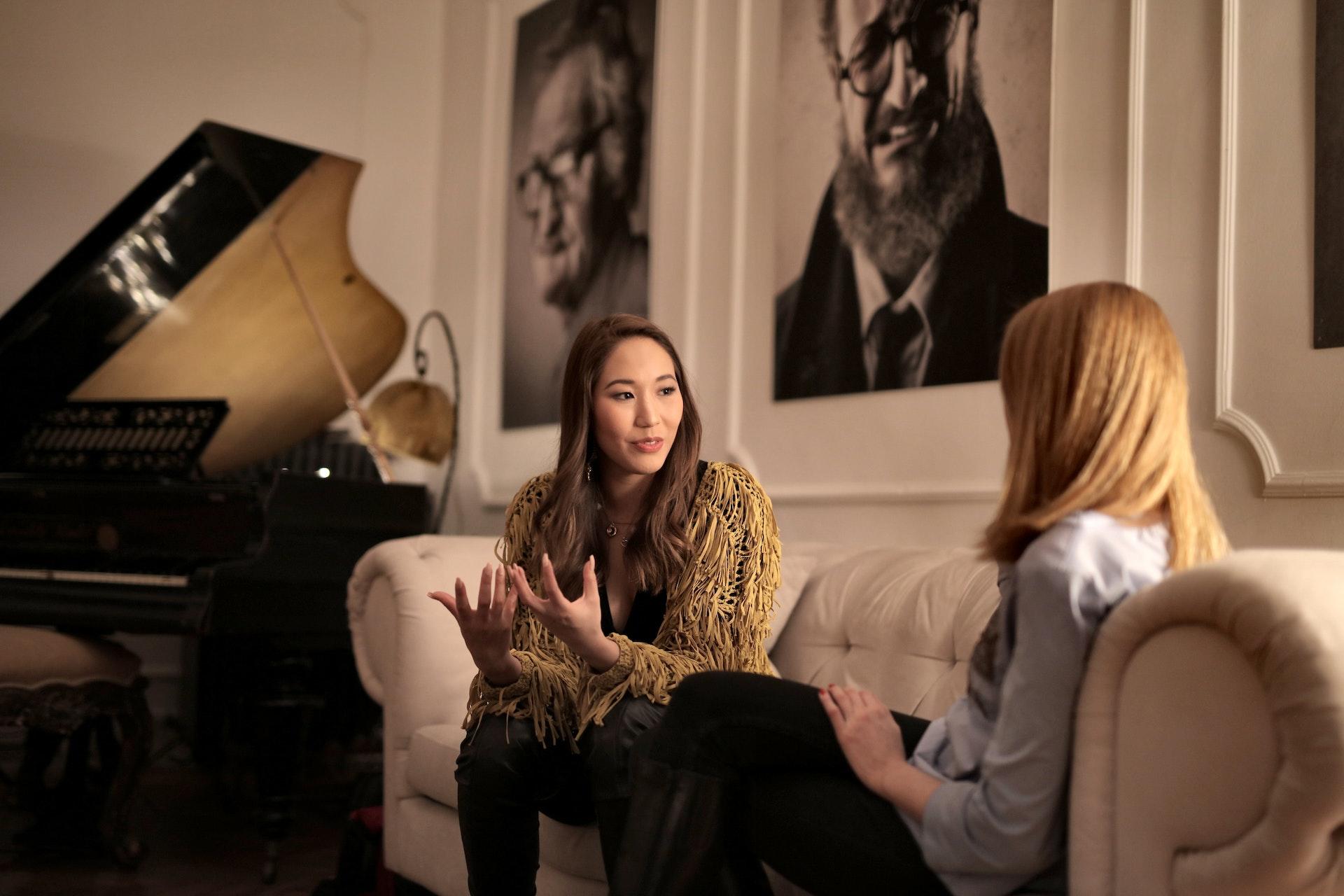 A piano teacher and student talk on a couch with the piano in the background.