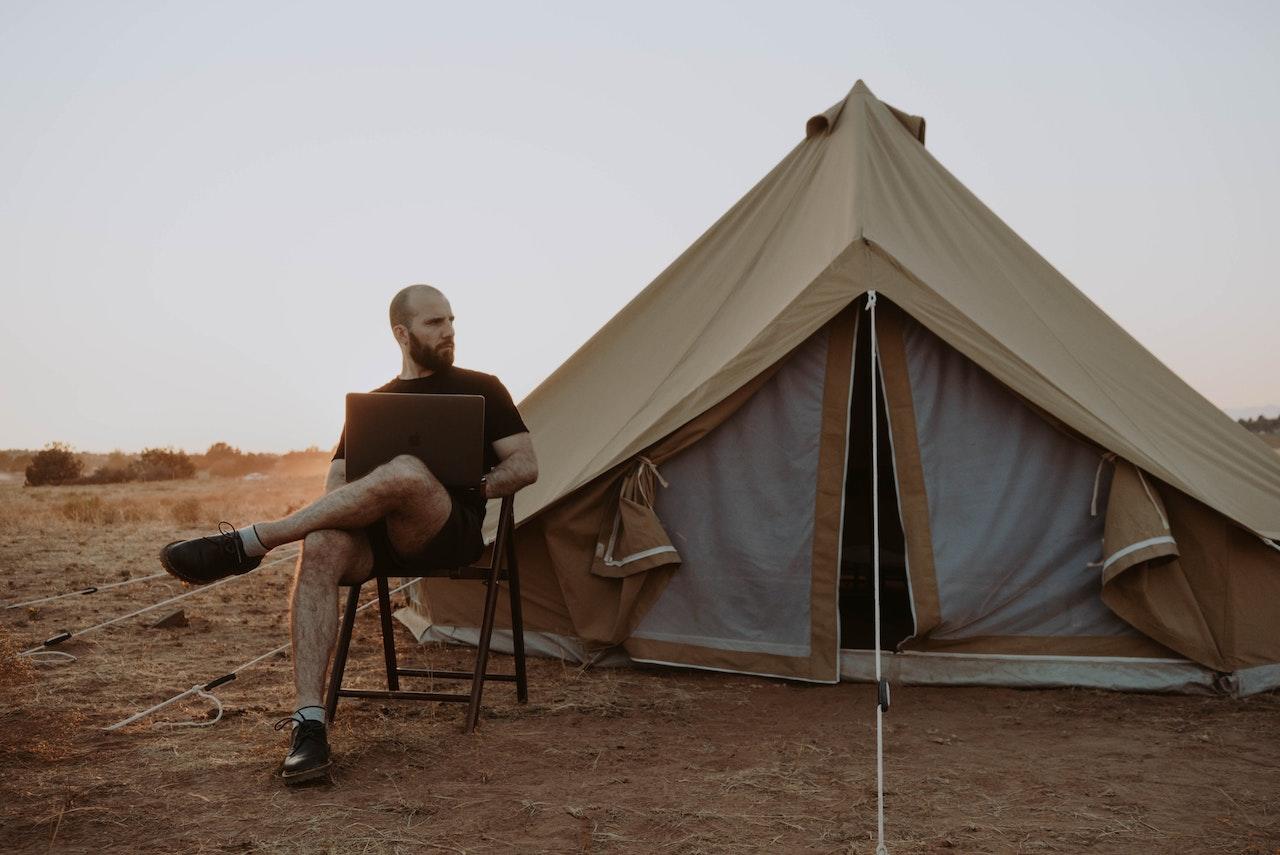 A man with a laptop sits in a chair near a tent in the desert.