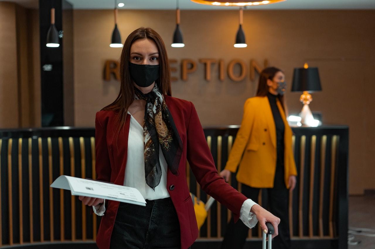 A woman in a red blazer standing in front of a hotel reception desk, holding her suitcase.