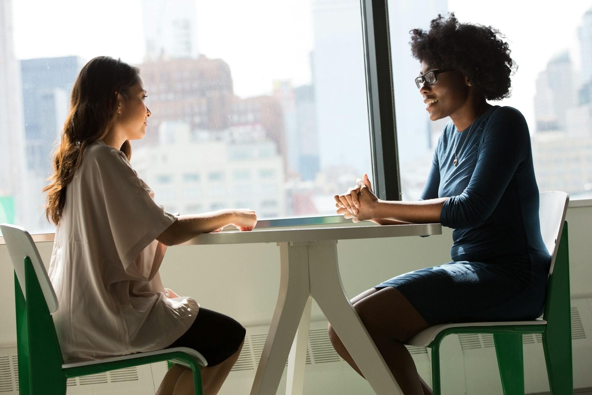 Two women talking across a table beside a large window