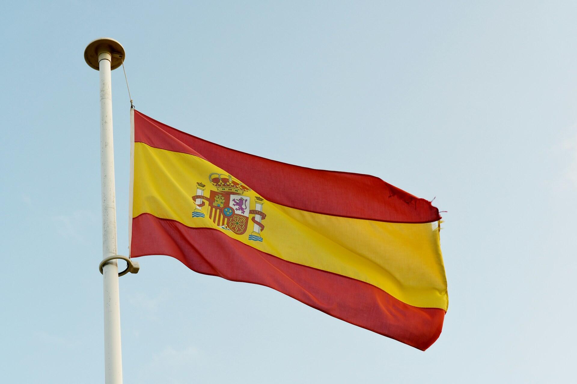 Spanish flag flying from a white flagpole against a pale blue sky