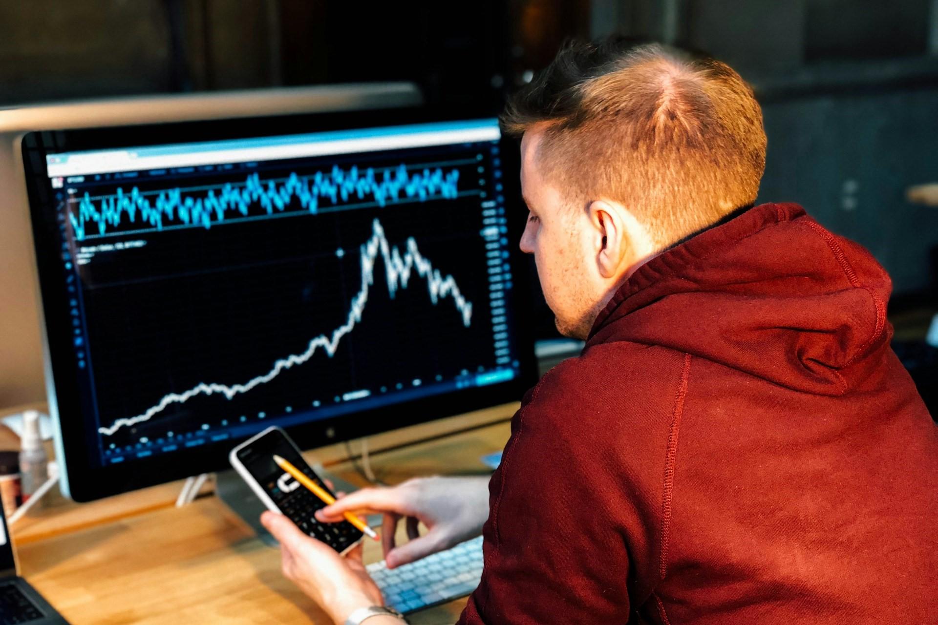 A man at a computer looking at stocks on a phone and the computer.
