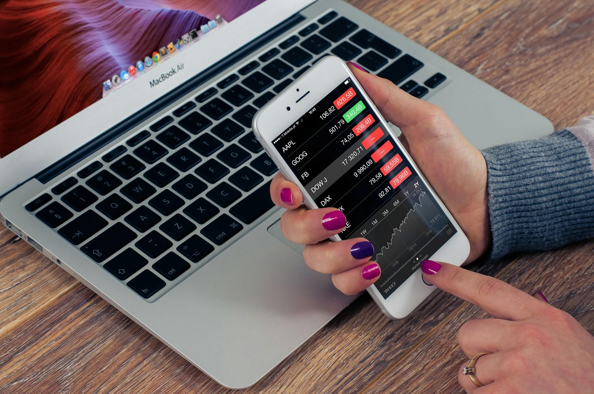 A hand holding a smartphone with stocks on it next to a Macbook.