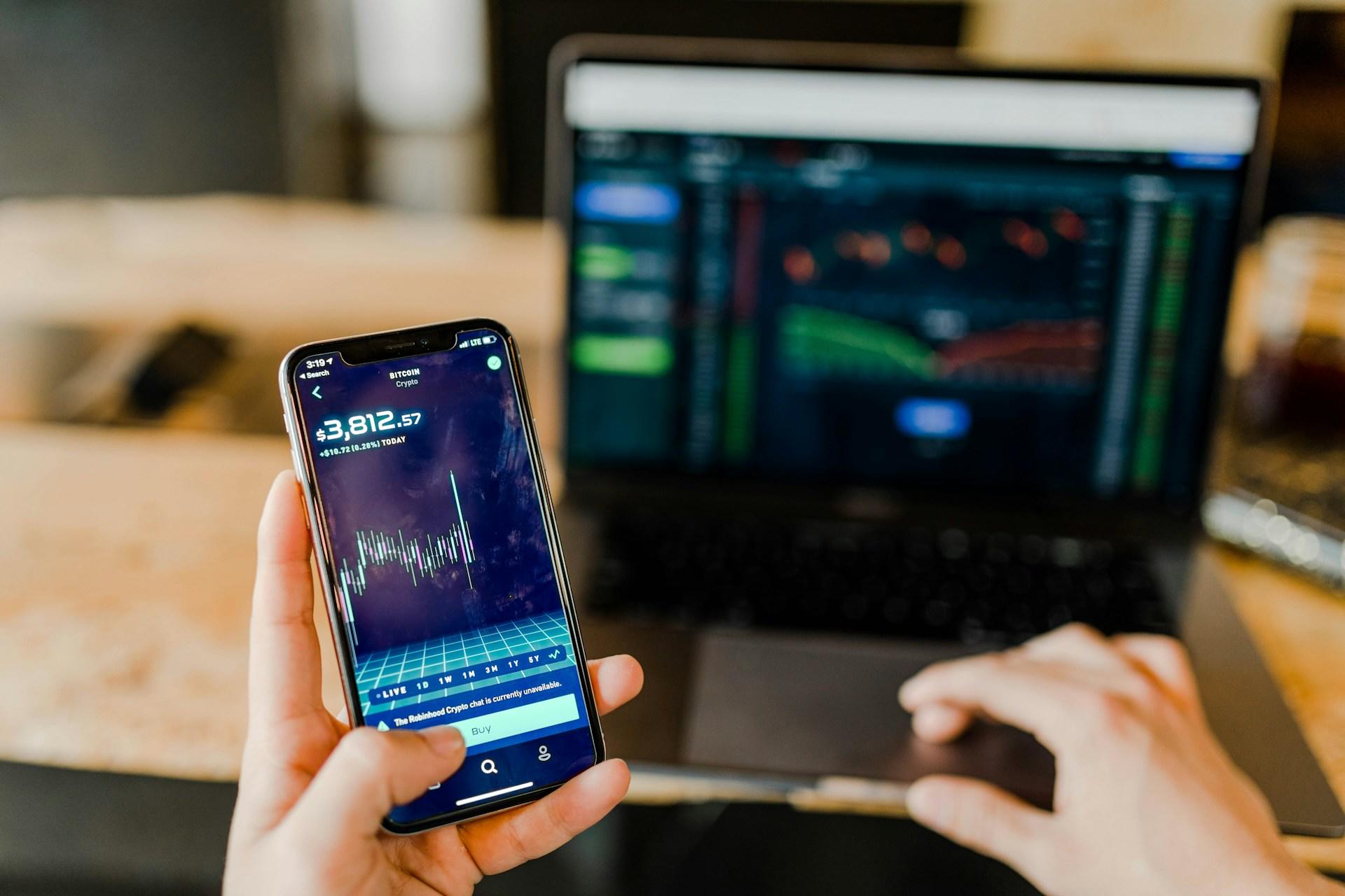 A hand holding a smartphone with a stock graph next to a computer with a graph on it.