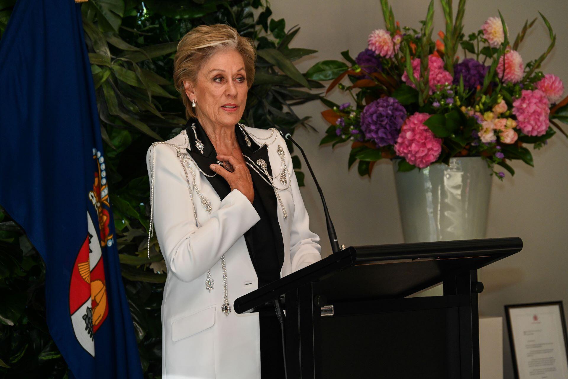 Dame Kiri Te Kanawa speaking at a podium with flowers in the background