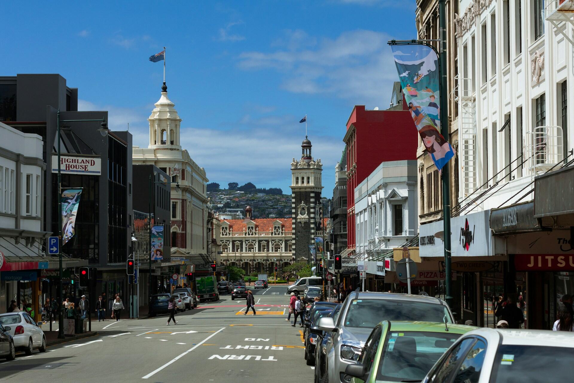 Central Dunedin street with historic buildings, traffic and the railway station clock tower in the distance.