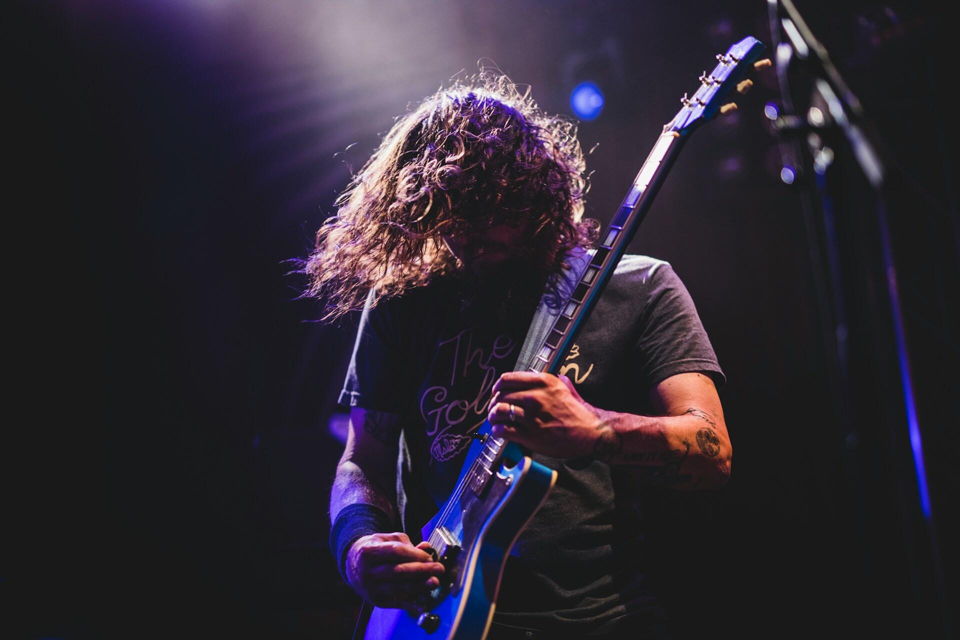 Guitarist with long curly hair playing an electric guitar under purple stage lights.