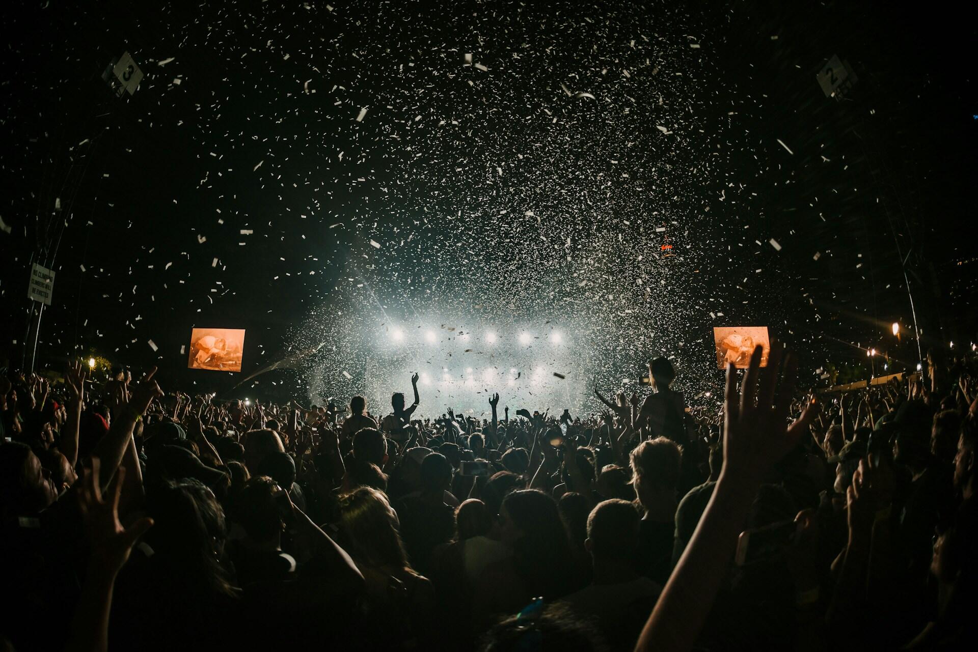 Large indoor concert crowd with raised hands and confetti falling in front of a brightly lit stage.