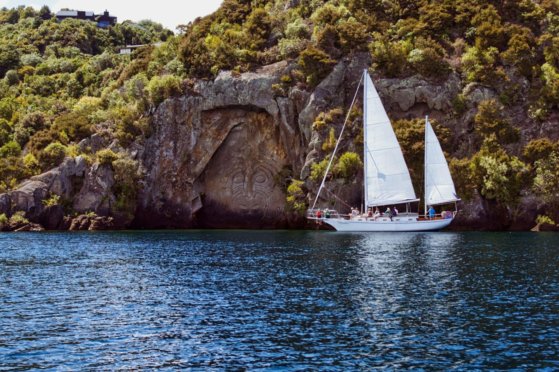 Sailing boat with white sails moving along a rocky coastline