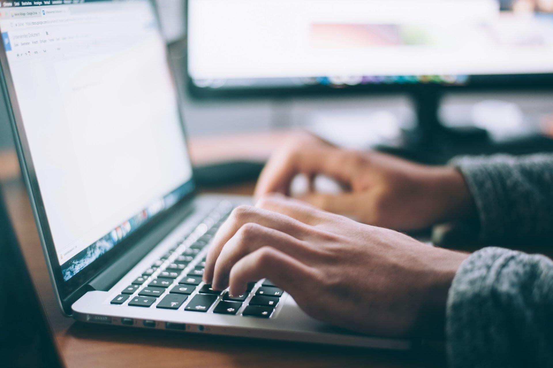 Hands typing on a laptop in front of computer screens